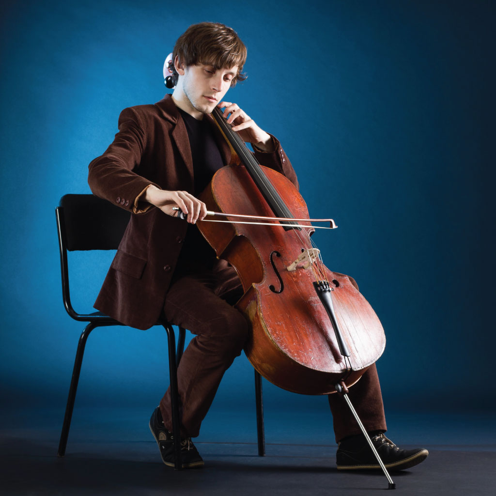 Young man playing a cello. He's on hgis own, sitting on a chair, with a blue background