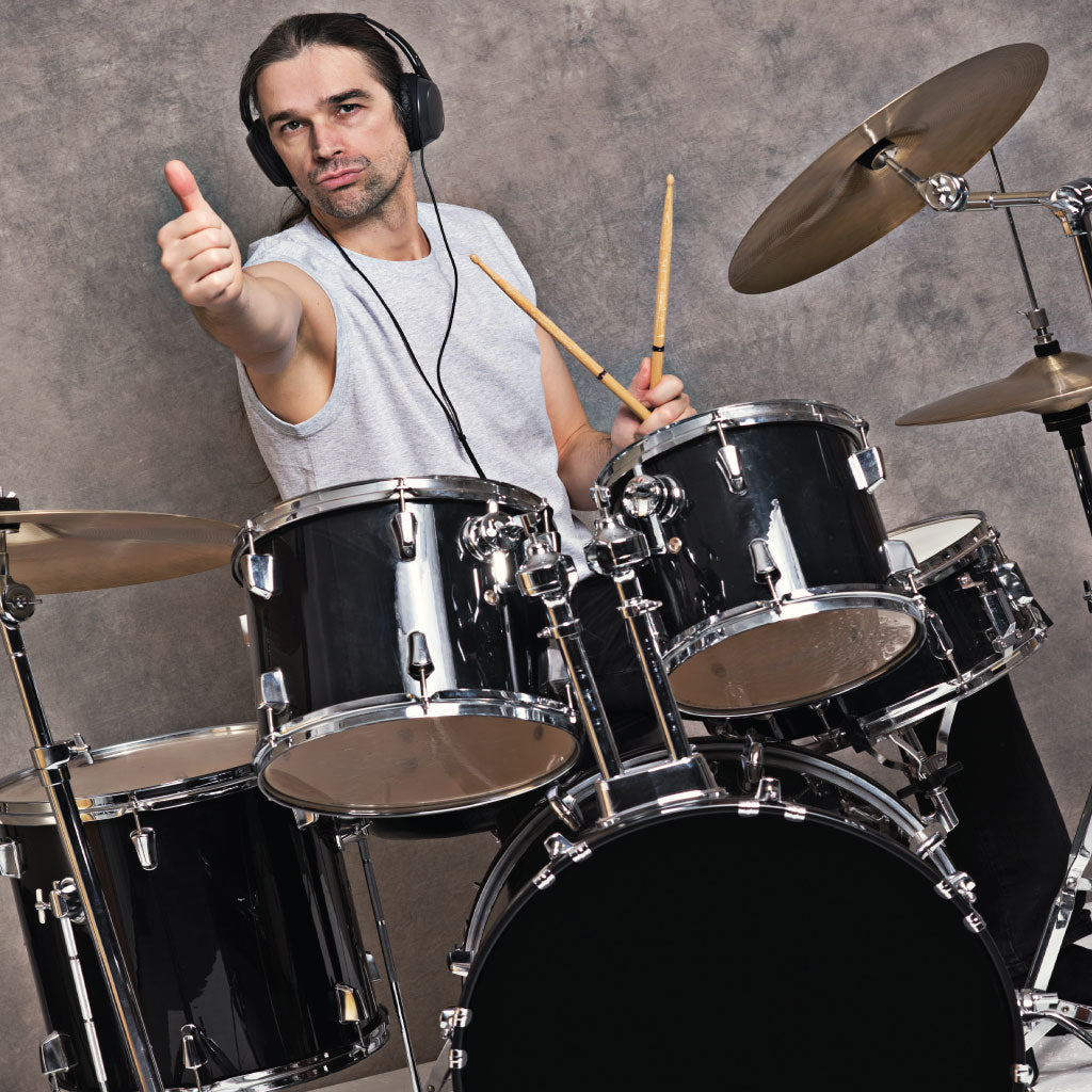 Man in white tee shirt sitting behind a black drum kit, holding drum sticks in one hand and giving a thumbs up with the other