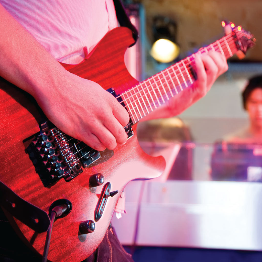 Cropped shot of a guitarist in the studio playing a superstrat style electric guitar. In the background there is someone behind glass in the control room looking on