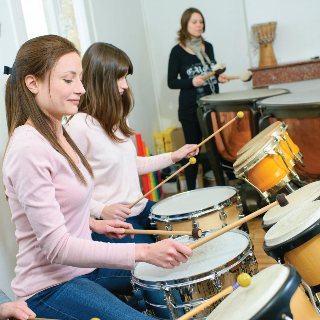 two young woman in the foreground playing percussion drums using mallets, with another in the background playing the kettle drums