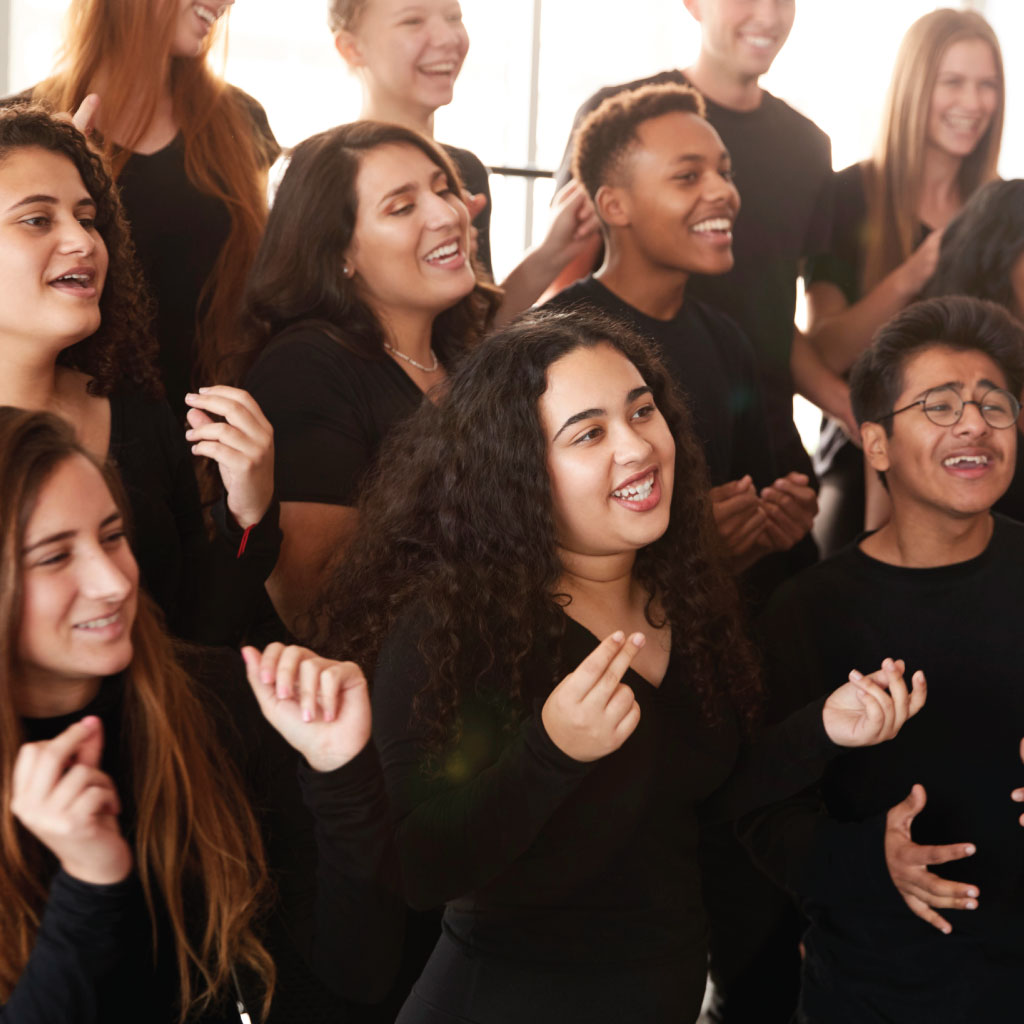 A cropped image of a choir, possibly students, all dressed in black, singing, snapping their fingers, and looking happy