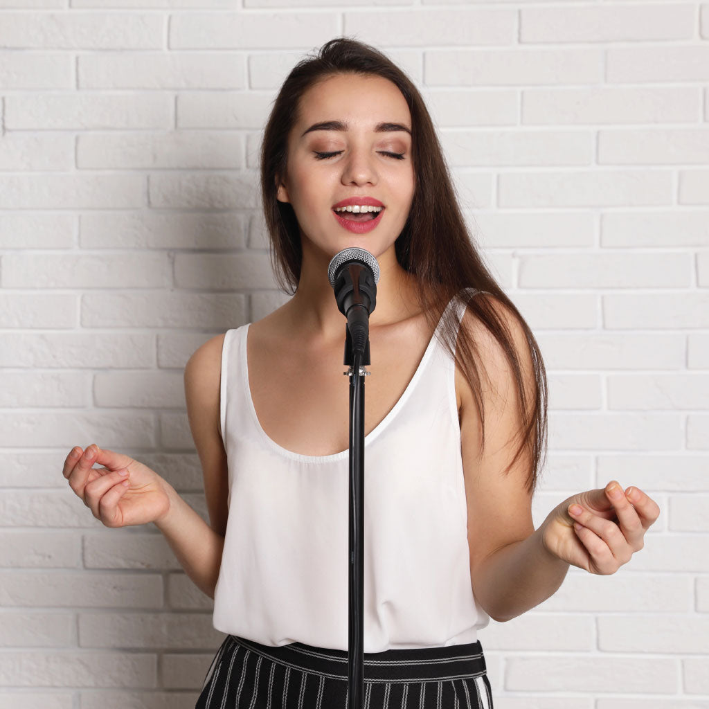 Young woman singing into a microphone. Background is a brick wall painted white